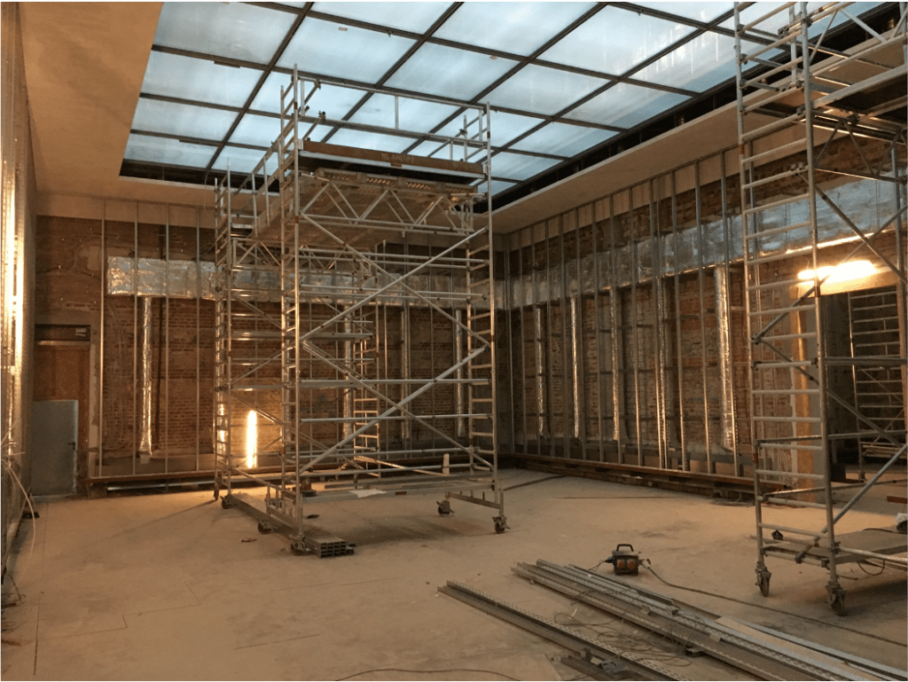 Eine große Baustelle im Innenbereich mit Metallgerüst, freiliegenden Wandrahmen, verstreuten Werkzeugen und einer Glasdecke, die natürliches Licht in den unfertigen Raum lässt.