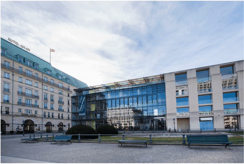 Neben dem historischen Hotel Adlon mit seinem Gründach steht ein modernes Glasgebäude; beide blicken auf einen offenen Platz mit leeren Bänken und gestutzten Büschen unter einem teilweise bewölkten Himmel.