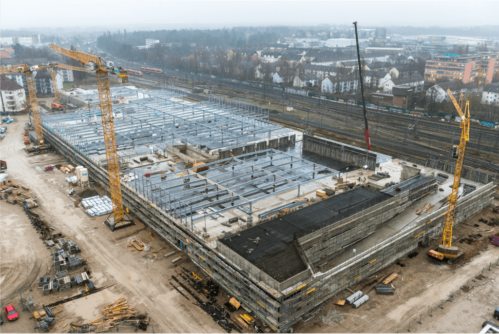Eine große Baustelle mit Kränen und teilweise fertiggestellten Strukturen, darunter Stahlgerüste und Betonwände. Um die Baustelle herum befinden sich Materialien, Ausrüstung und nahegelegene Bahngleise. Im Hintergrund sind Gebäude und Bäume zu sehen.