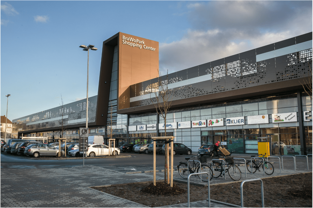 Ein modernes Einkaufszentrum mit einem großen Schild mit der Aufschrift „BraWoPark Shopping Center“ und geparkten Autos, Fahrrädern und Ladenschildern im Erdgeschoss unter einem teilweise bewölkten Himmel.