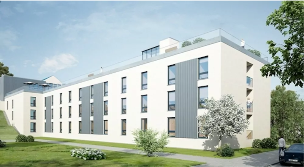 Ein modernes, dreistöckiges Apartmentgebäude mit großen Fenstern und vertikalen grauen Paneelen, umgeben von grünem Gras, kleinen Bäumen und Sträuchern unter einem blauen Himmel.