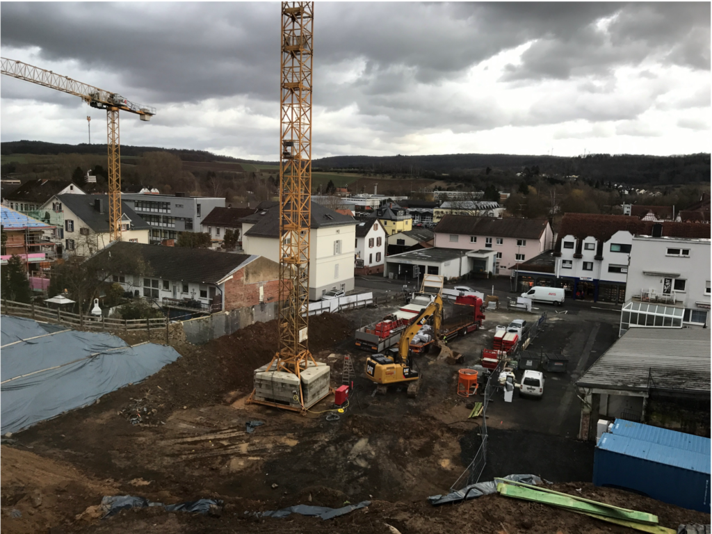 Vor Wohn- und Geschäftshäusern ist unter bewölktem Himmel eine Baustelle mit Kränen, Baggern und Geräten zu sehen. Im Hintergrund sind Häuser, Straßen und eine hügelige Landschaft zu sehen.
