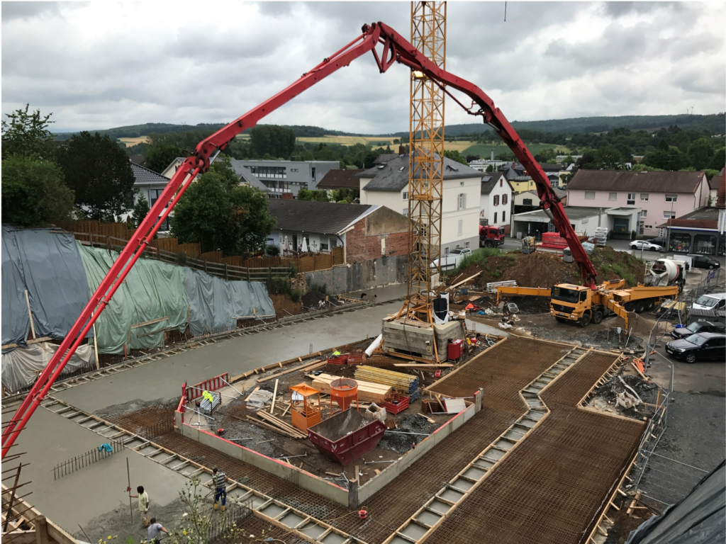 Eine Baustelle: Arbeiter gießen Beton mit einem großen roten Betonpumpenwagen. Bewehrungsgitter und Baumaterialien sind zu sehen, umgeben von Häusern und Bäumen unter einem bewölkten Himmel.