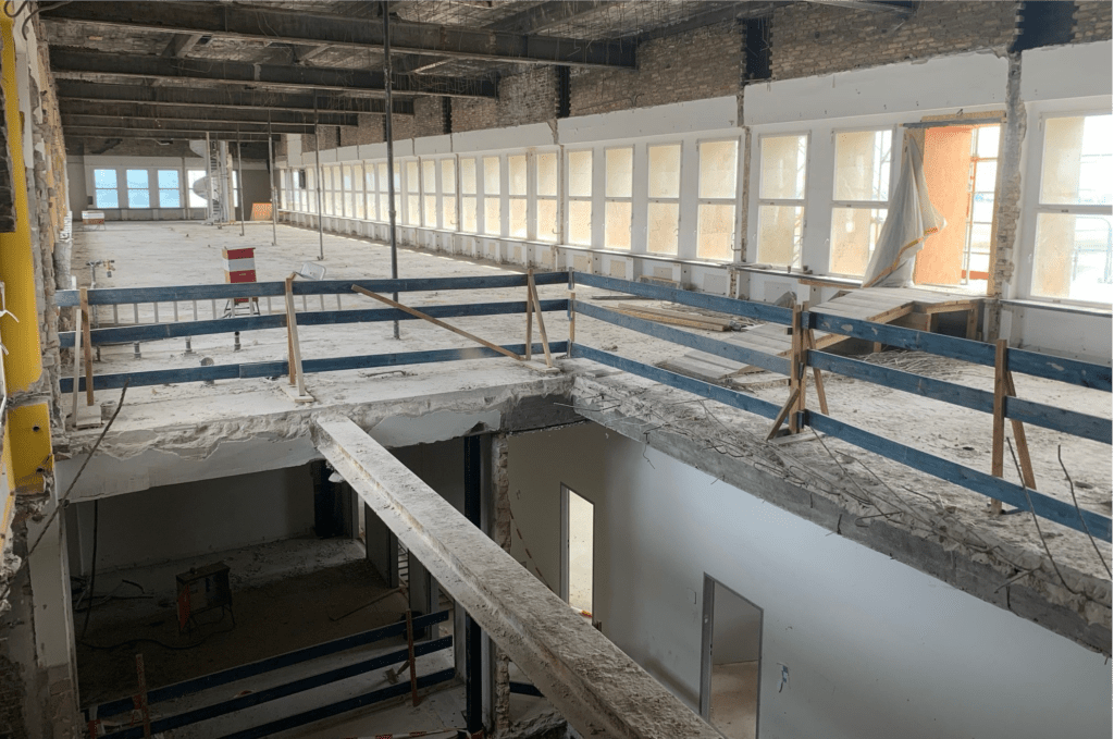 Ein großes Gebäude wird gerade renoviert. Es weist freiliegende Balken, unfertige Wände und Bauzäune auf. Sonnenlicht fällt durch die Fensterreihen, und Schutt liegt verstreut auf beiden Etagen.