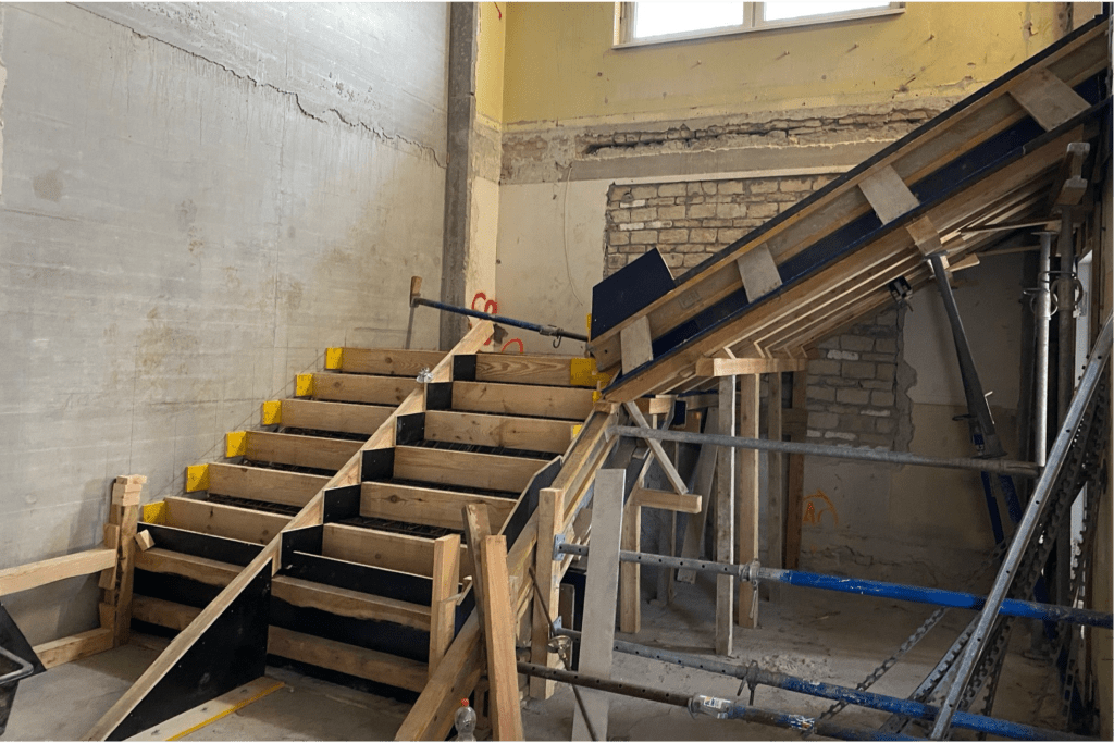 Eine Baustelle mit einem Holzrahmen für den Treppenbau, Gerüsten und freiliegenden Ziegel- und Betonwänden in einem teilweise fertiggestellten Raum. Licht fällt durch ein Fenster darüber ein.
