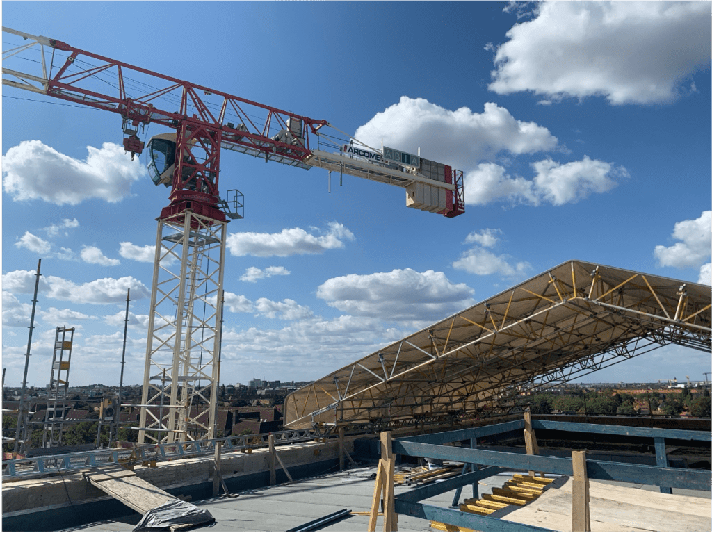 Eine Baustelle unter blauem Himmel mit vereinzelten Wolken, mit einem großen rot-weißen Kran und einer teilweise errichteten Dachkonstruktion, auf der Gerüste und Baumaterialien sichtbar sind.