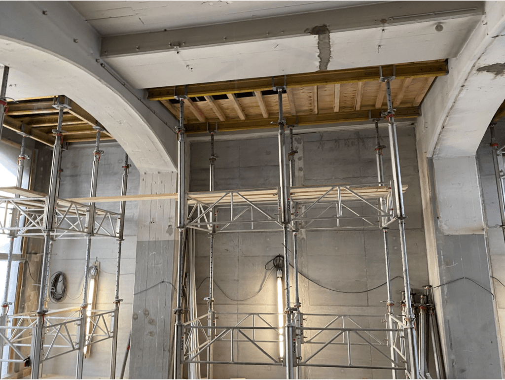 In einem im Bau befindlichen Gebäude steht ein Metallgerüst an einer hohen Betonwand. An der Decke sind Holzbretter und freiliegende Balken sichtbar. Der Arbeitsbereich wird durch eine Beleuchtung erhellt.