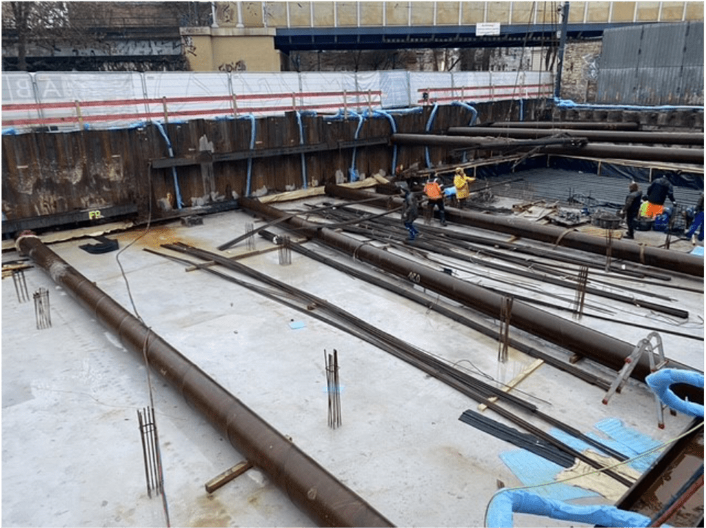Baustelle mit großen Stahlträgern auf einem Betonfundament. Mehrere Arbeiter in Schutzausrüstung stehen in der Nähe der Ausrüstung. Stützmauern und Schutzbarrieren säumen die Baustelle.