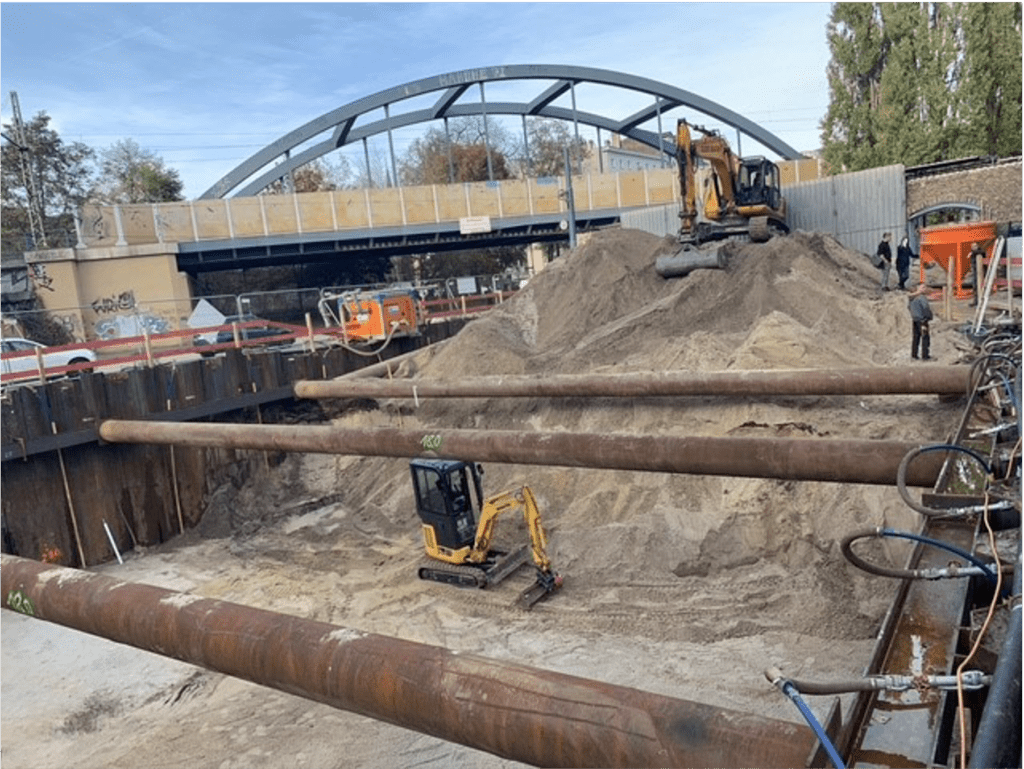 Eine Baustelle mit zwei Baggern, die an einer tiefen, mit Sand gefüllten Grube arbeiten. Große Stahlrohre spannen sich über die Grube, im Hintergrund ist eine Brücke mit einem Metallbogen zu sehen. Baufahrzeuge und Zäune umgeben das Gelände.
