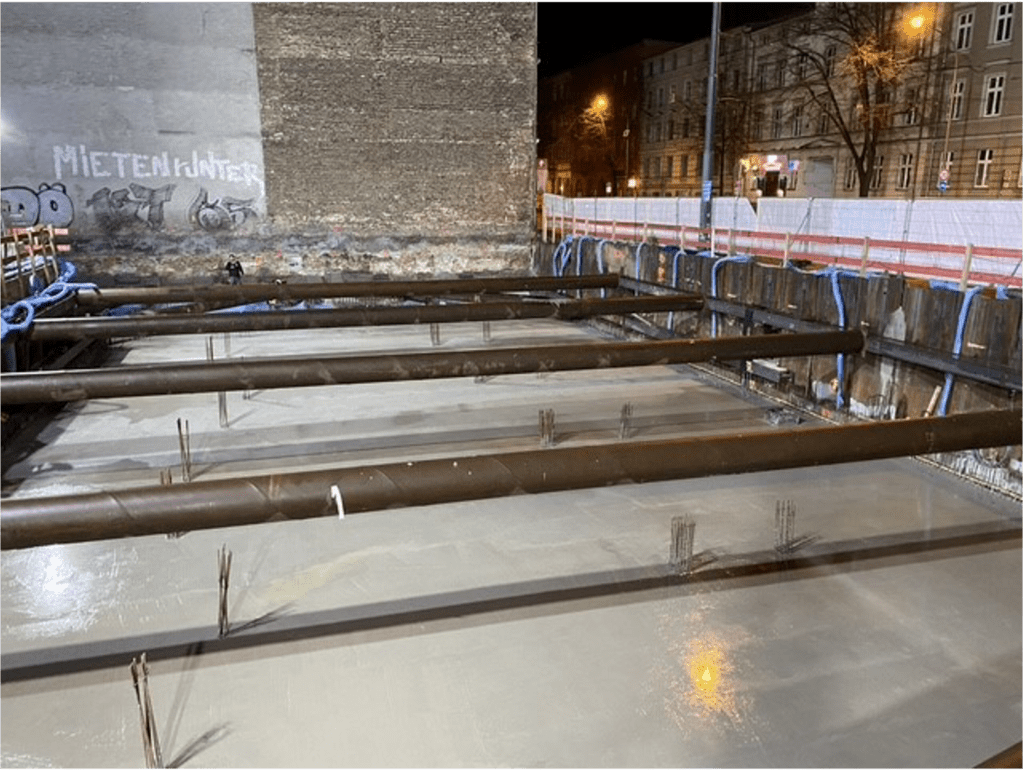 Eine nächtliche Baustelle zeigt ein Betonfundament mit großen Stahlträgern, die das Gelände überspannen. Im Hintergrund sind Straßenlaternen und Gebäude zu sehen. An der Wand steht das Graffiti „MIETEN WAHNSINN.“.