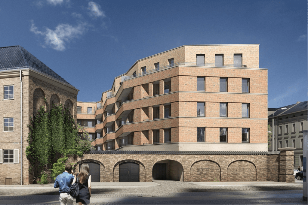 Ein modernes, mehrstöckiges Backsteingebäude mit großen Fenstern und Balkonen steht neben einem älteren, mit Efeu bewachsenen Backsteingebäude. Zwei Menschen gehen unter einem strahlend blauen Himmel auf der Kopfsteinpflasterstraße vor den Gebäuden.