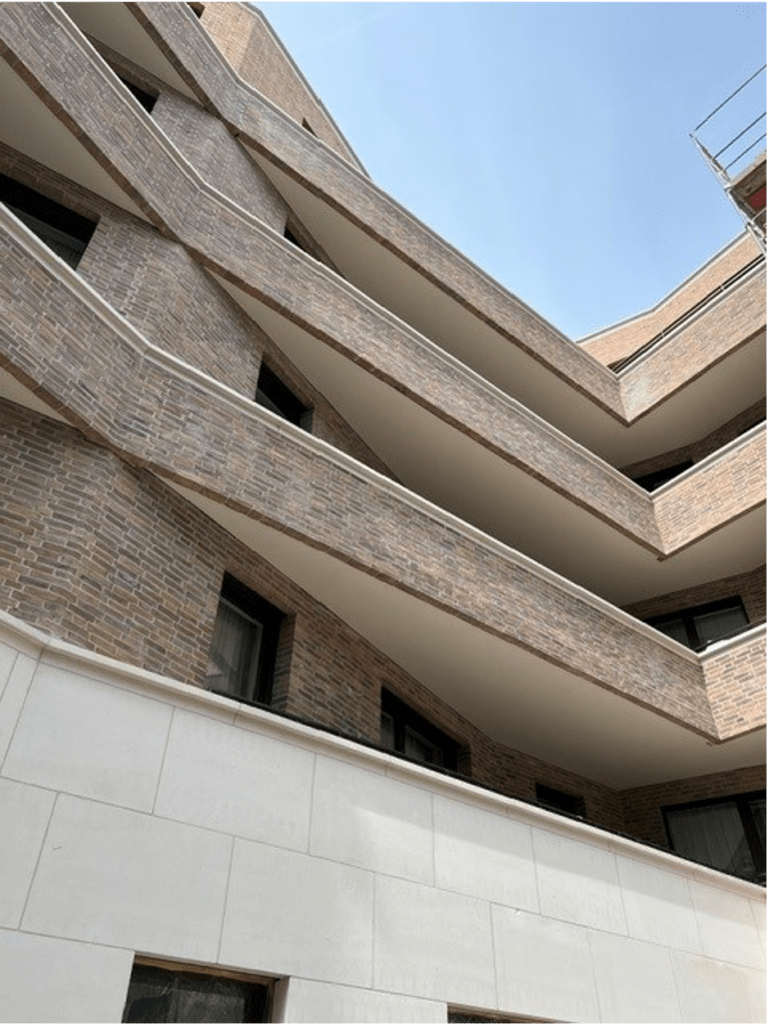 Ein modernes Gebäude mit eckigen Balkonen und braunem Mauerwerk, von unten betrachtet vor einem klaren blauen Himmel. Die Struktur zeichnet sich durch geometrische Linien und kontrastierende weiße Unterwände aus.