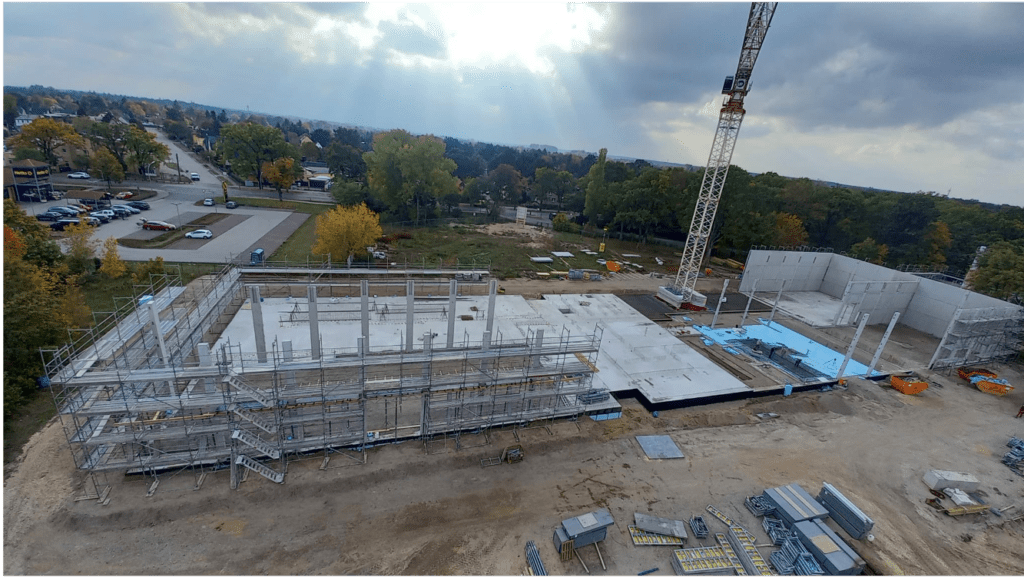 Eine Baustelle mit der Fundamentierung und dem Rahmen eines großen Gebäudes im Gange, ein Kran in der Nähe, überall verstreute Materialien und Bäume und Straßen im Hintergrund unter einem bewölkten Himmel mit Sonnenstrahlen.