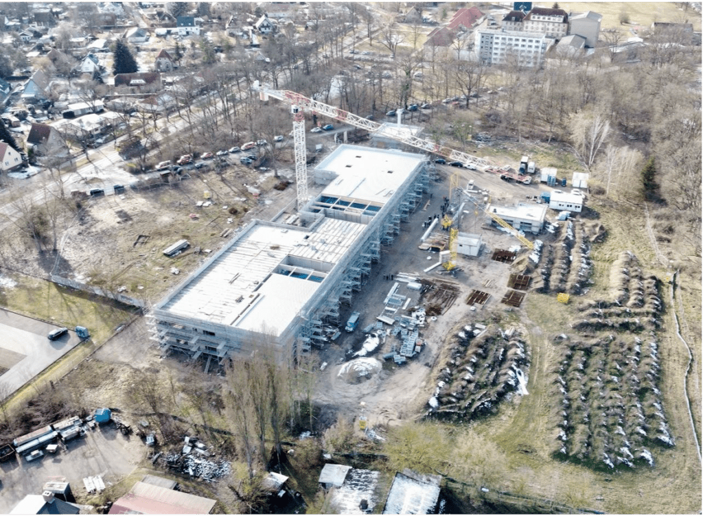 Luftaufnahme einer Baustelle mit einem großen rechteckigen Gebäude, einem Kran, Baumaterialien und Geräten. In der Umgebung befinden sich Bäume, bepflanzte Reihen und Wohnhäuser. Auf dem Boden sind Schneeflecken sichtbar.