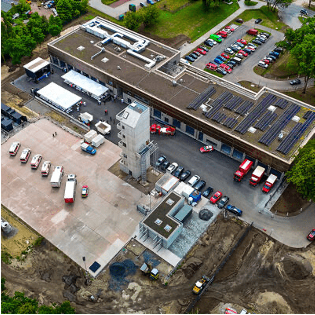 Luftaufnahme eines Feuerwachenkomplexes mit mehreren draußen geparkten Feuerwehrautos, einem Übungsturm, Solarmodulen auf dem Dach, Parkplätzen mit Autos und umgebender Grünanlage.