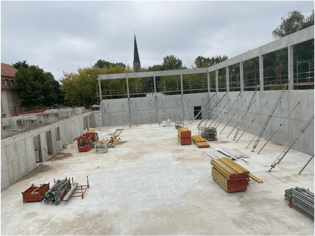 Eine Baustelle mit hohen Betonmauern, Metallträgern und Stapeln von Baumaterialien. Im Hintergrund sind Bäume und ein Kirchturm unter einem bewölkten Himmel zu sehen.
