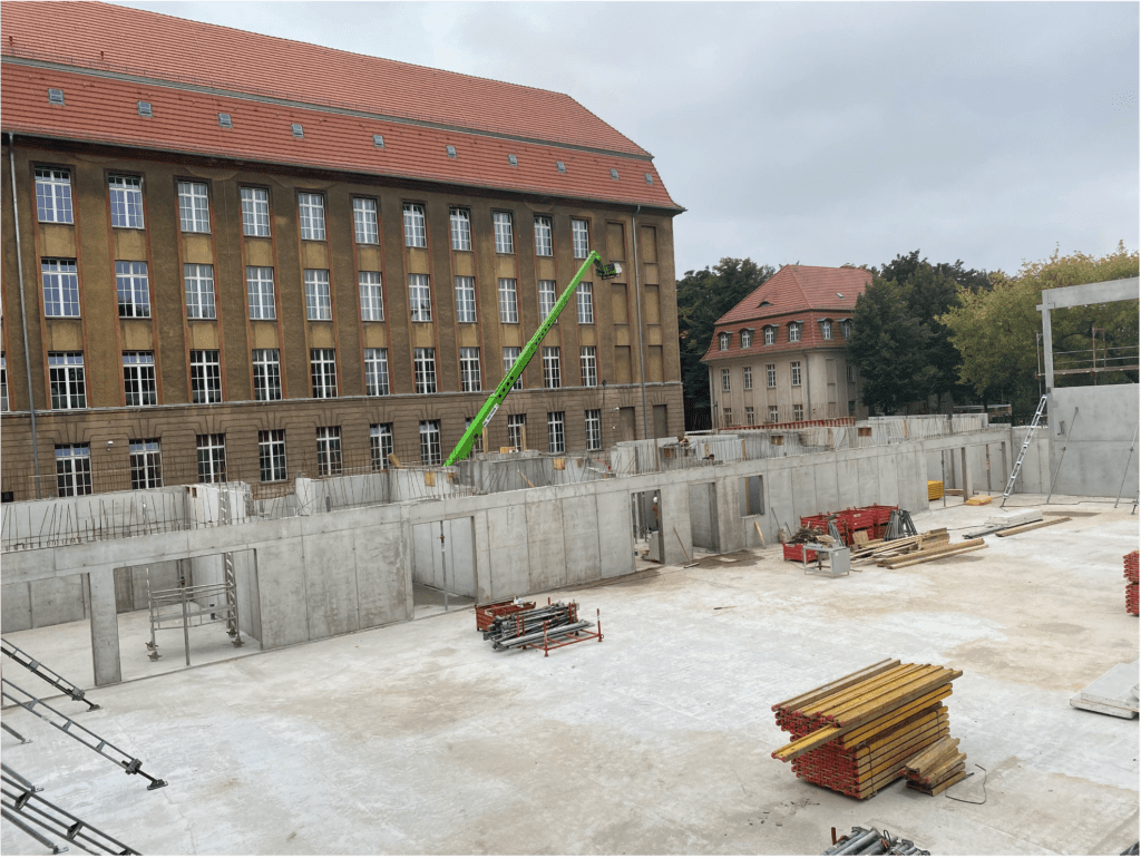 Eine Baustelle mit Betonwänden und Baumaterialien im Vordergrund, daneben ein großes, mehrstöckiges Backsteingebäude mit vielen Fenstern und einem roten Ziegeldach. Ein grüner Kran ragt bis zum Gebäude hinauf.