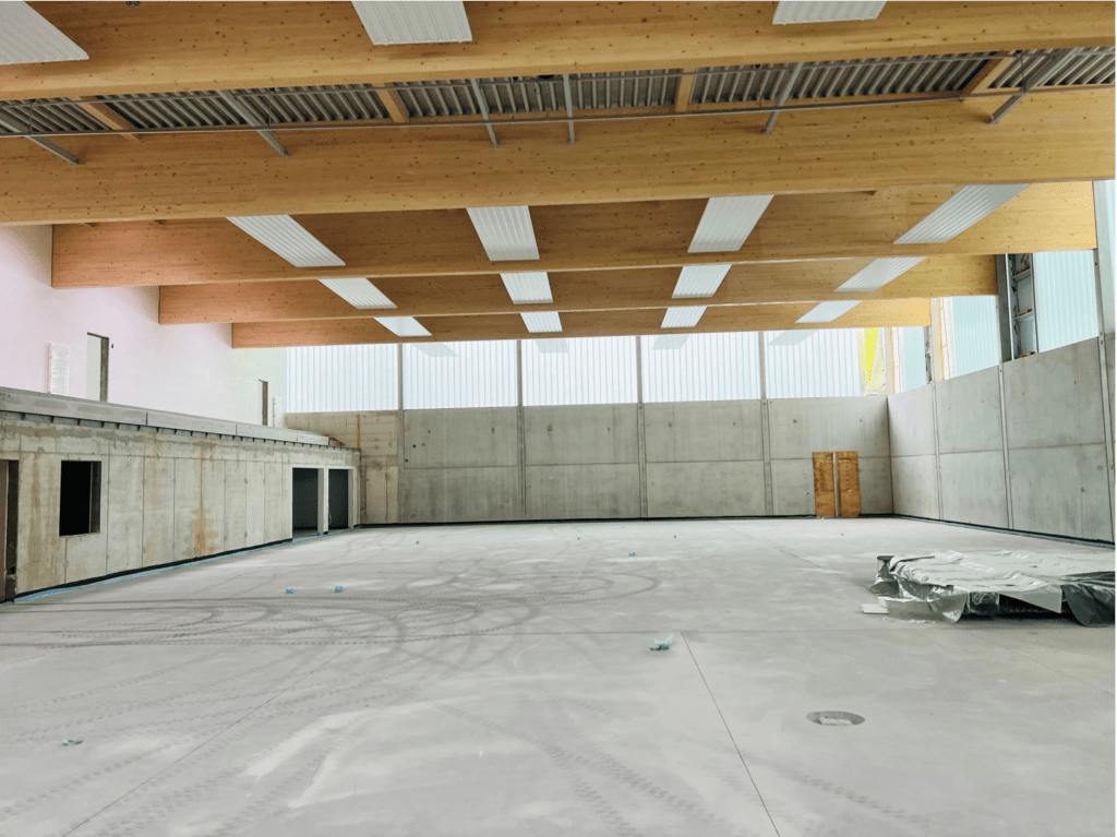 Eine leere, unfertige Sporthalle mit Betonboden und -wänden, freiliegenden Holzbalken an der Decke und großen Fenstern, die Tageslicht hereinlassen. Baumaterialien liegen verstreut auf dem Boden.