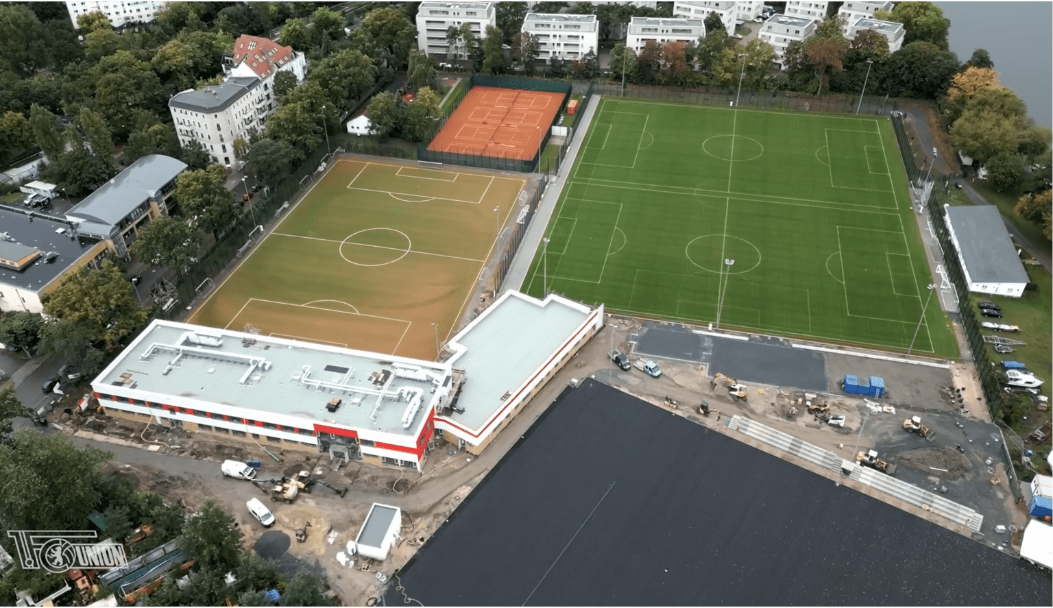 Luftaufnahme eines Sportkomplexes mit einem grünen Fußballfeld, einem braunen Mehrzweckfeld, einem Tennisplatz, einem weiß-roten Gebäude sowie umliegenden Bäumen und Wohnhäusern. In der Nähe des Eingangs sind einige Bauarbeiten zu sehen.