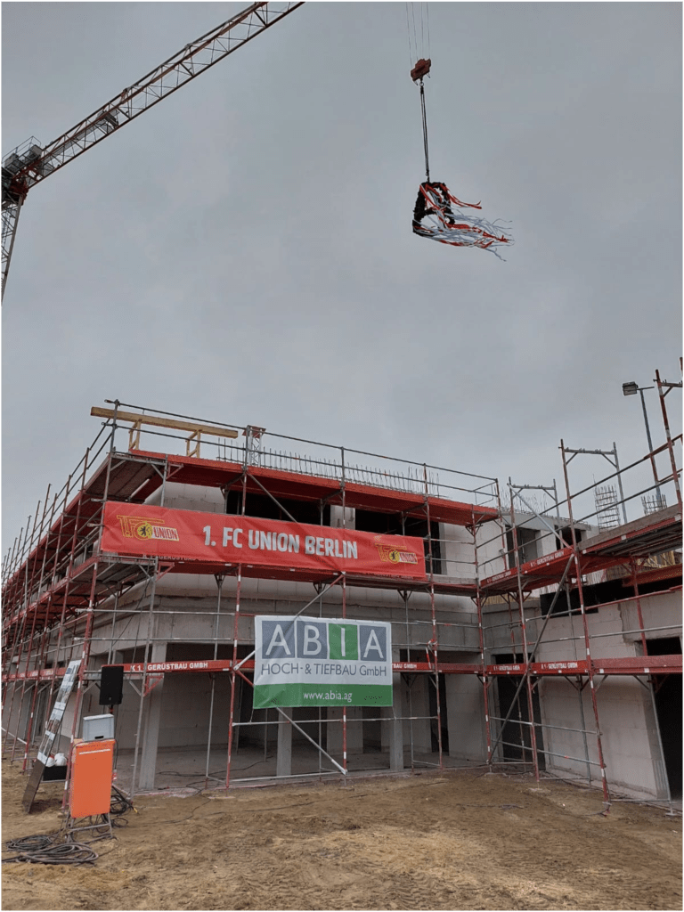 Eine Baustelle mit Gerüsten und einem Kran, der Material hebt. Das Gebäude ist teilweise fertiggestellt, auf Transparenten sind „1. FC UNION BERLIN“ und „ABA Hoch- & Tiefbau GmbH“ zu sehen. Der Himmel ist bedeckt.