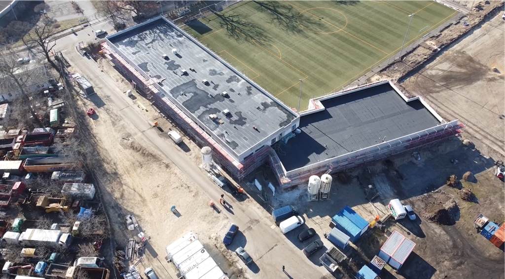 Luftaufnahme einer Baustelle mit Gebäuden im Umbau neben einem grünen Kunstrasen-Sportplatz. Fahrzeuge, Baumaschinen und Lagercontainer sind rund um die Baustelle zu sehen.
