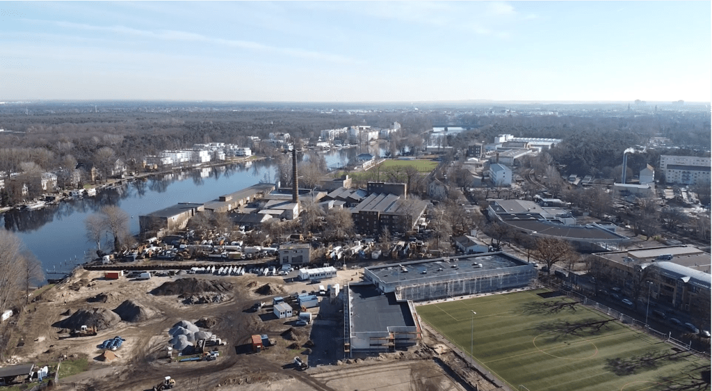 Luftaufnahme eines städtischen Flussgebiets mit Gebäuden, Baustellen, einem Fußballplatz, geparkten Fahrzeugen und einem von Bäumen umgebenen Fluss. Im Hintergrund sind Wohn- und Geschäftsgebäude zu sehen.