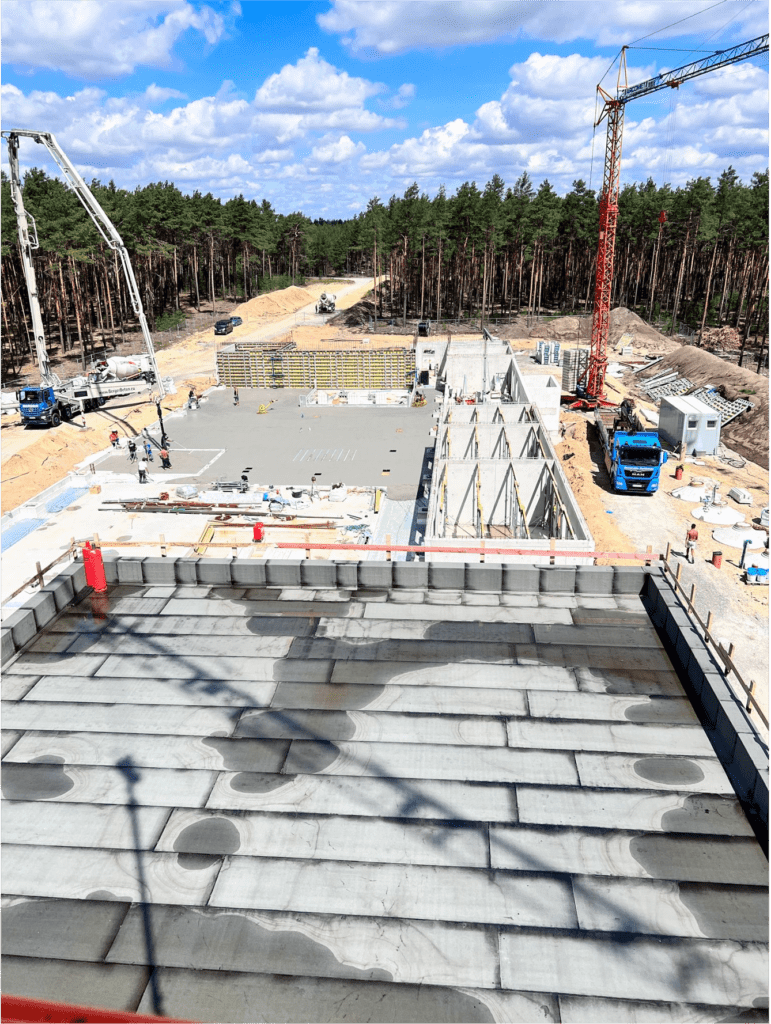 Baustelle von oben gesehen. Betonfundamente, Baumaterialien, Kräne, Lastwagen und Wald im Hintergrund unter leicht bewölktem Himmel sind zu sehen. Arbeiter und Baumaschinen sind auf der Baustelle zu sehen.