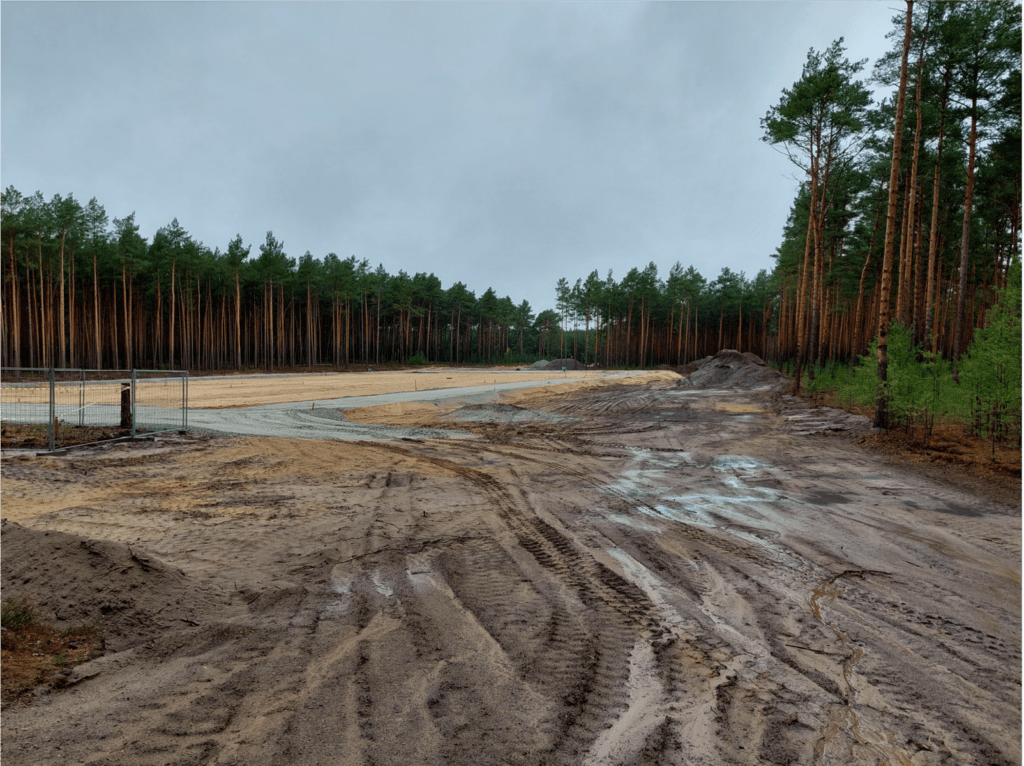Eine schlammige Baustelle mit tiefen Reifenspuren, umgeben von hohen Kiefern unter einem bewölkten Himmel. Links steht ein Metallzaun, in der Ferne sind Erdhaufen zu sehen.