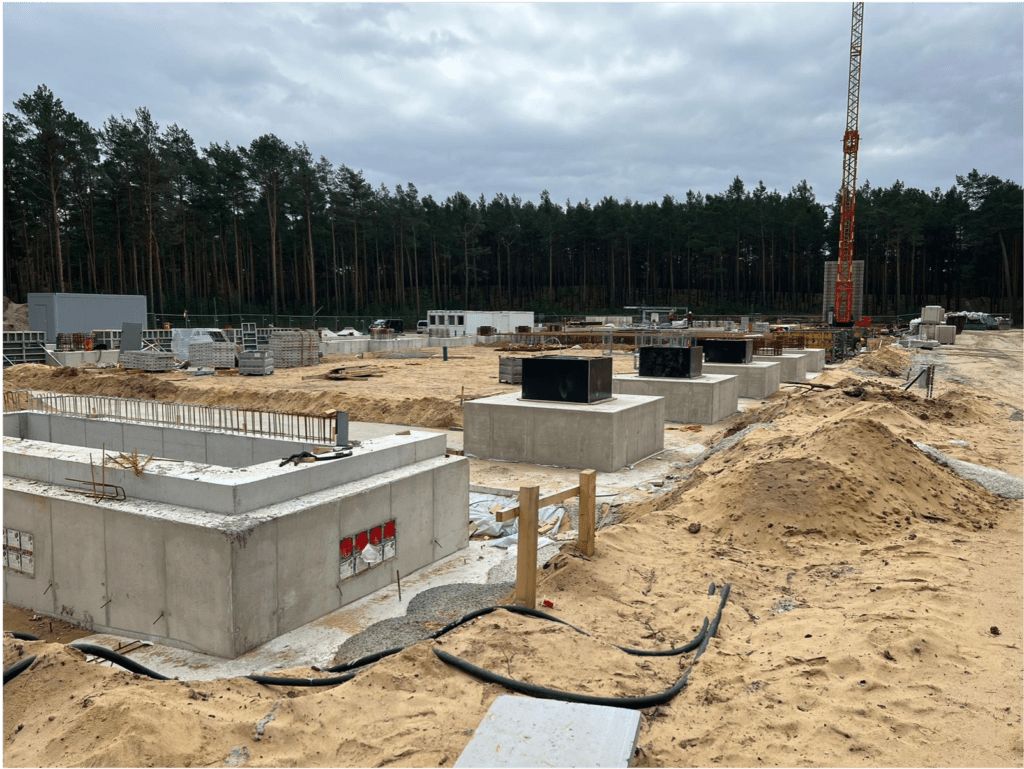 Eine Baustelle mit mehreren großen Betonfundamenten im Vordergrund, Stromkabeln auf sandigem Boden und einem Kran und Waldgebiet im Hintergrund unter einem bewölkten Himmel.