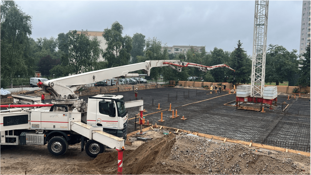 Ein Betonpumpenwagen fährt seinen langen Ausleger über eine Baustelle aus, wo Arbeiter an einem bewölkten Tag das Fundament mit Bewehrungsgittern und Holzschalungen vorbereiten. Im Hintergrund sind Bäume und geparkte Autos zu sehen.