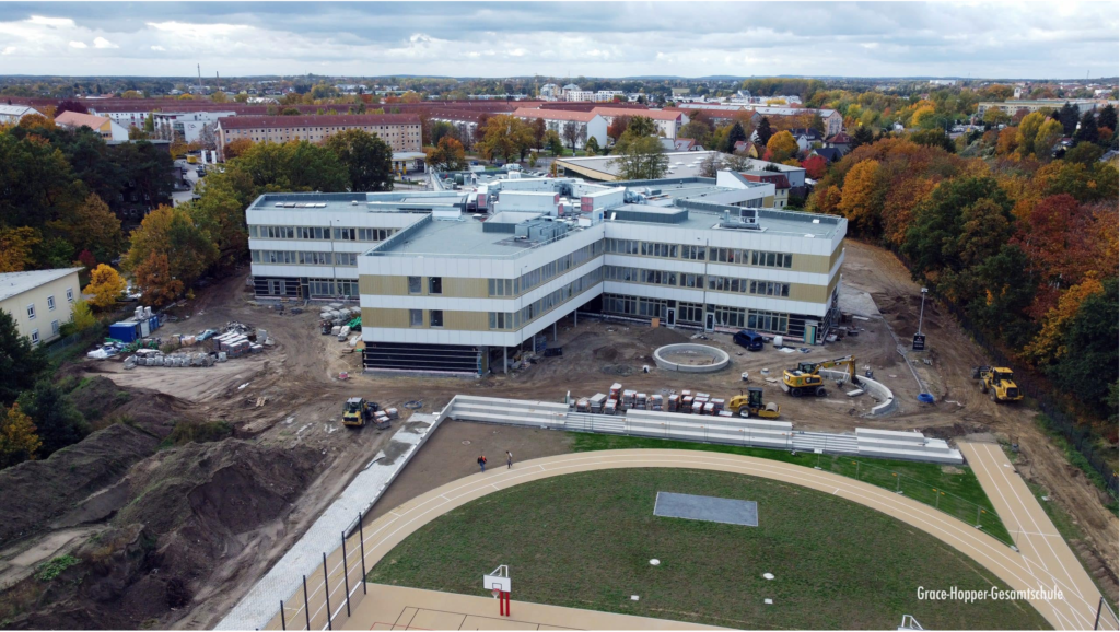 Luftaufnahme eines im Bau befindlichen modernen Schulgebäudes, umgeben von Baufahrzeugen und -materialien, mit einer fertiggestellten Sportbahn und Herbstbäumen im Hintergrund.