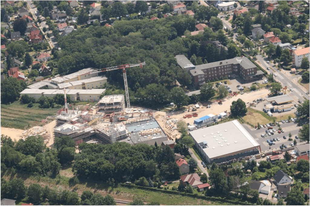 Luftaufnahme einer Baustelle mit einem Kran, umgeben von Bäumen und mehreren Gebäuden, in einem Vorortgebiet mit Straßen, Häusern und Grünflächen.