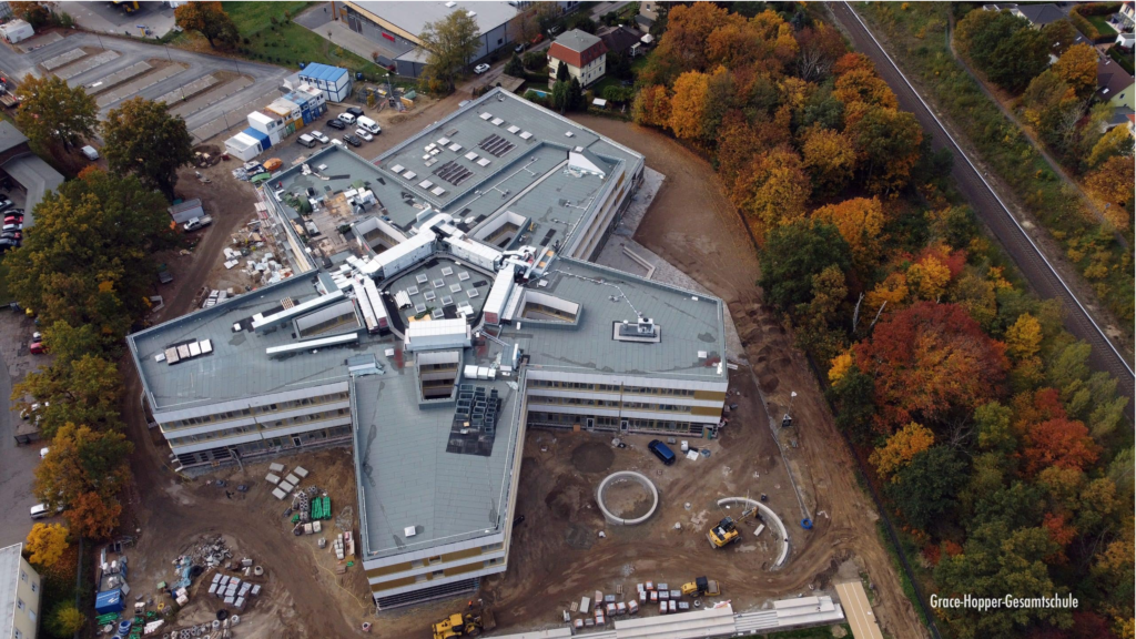 Luftaufnahme eines großen, sternförmigen Gebäudes im Bau, umgeben von Baumaterialien, Geräten, Herbstbäumen, nahegelegenen Straßen und Eisenbahnschienen.