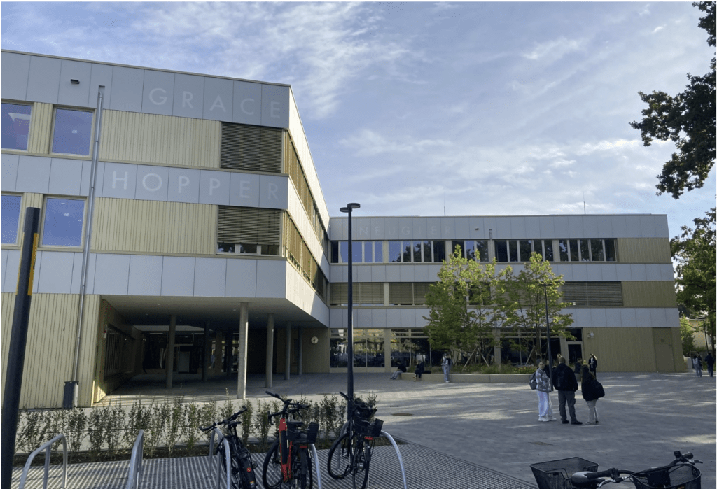 Ein modernes Gebäude mit der Aufschrift „GRACE HOPPER“ an der Fassade, großen Fenstern und Holzpaneelen. Davor parken Fahrräder, und mehrere Menschen gehen oder stehen in der Nähe. Bäume und blauer Himmel sind zu sehen.