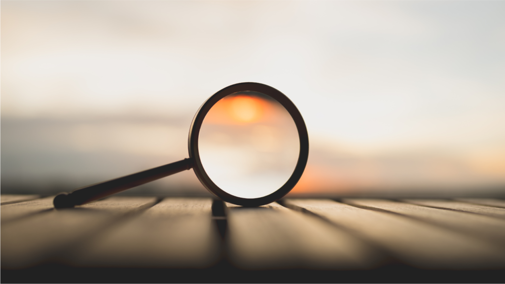 Eine Lupe liegt auf einer Holzoberfläche mit dem Sonnenuntergang im Hintergrund und erzeugt einen warmen, verschwommenen Schein, der durch und um die Linse herum sichtbar ist.