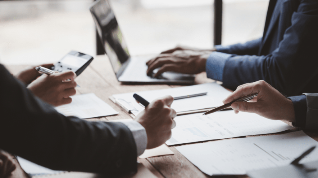 Drei Personen sitzen an einem Tisch, arbeiten an Laptops und Dokumenten und besprechen Finanzdiagramme und -daten. Eine Person hält ein Smartphone und eine andere zeigt mit einem Stift auf ein Blatt Papier.