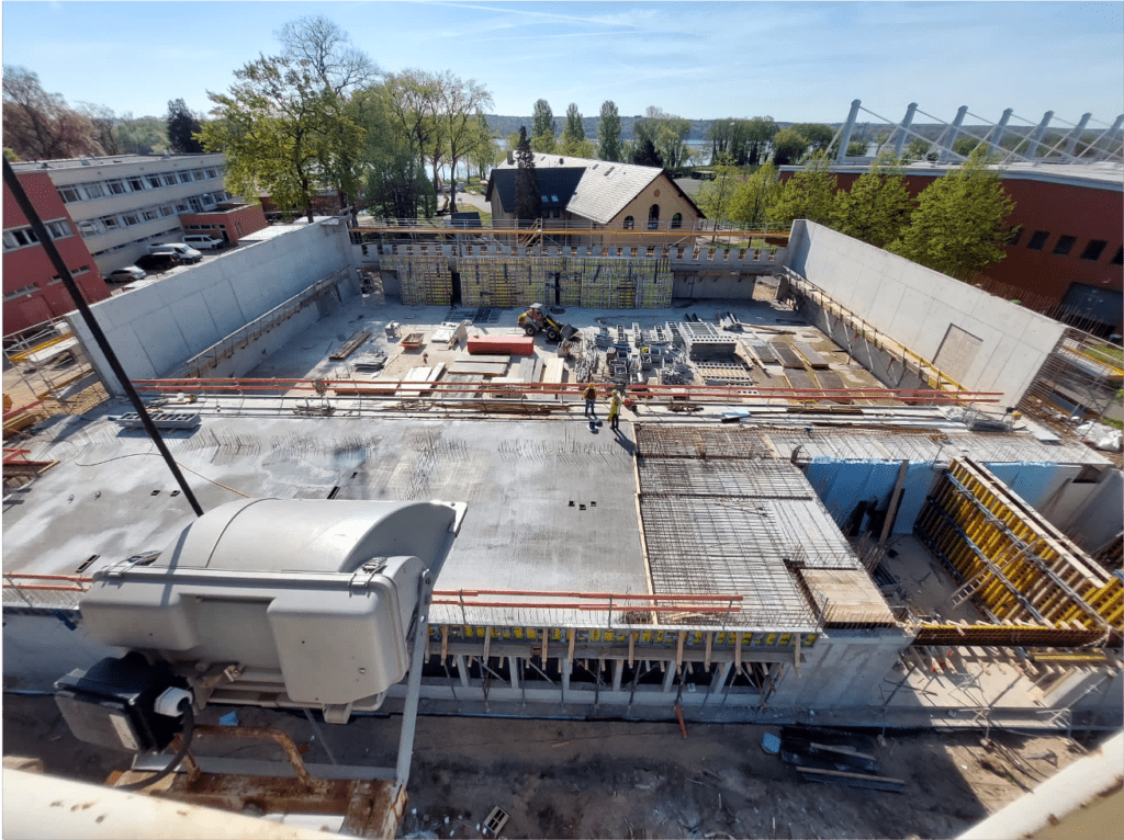 Luftaufnahme einer großen Baustelle mit Arbeitern, Baumaterialien und Maschinen, umgeben von hohen Betonmauern und nahe gelegenen Gebäuden, mit Bäumen und blauem Himmel im Hintergrund.