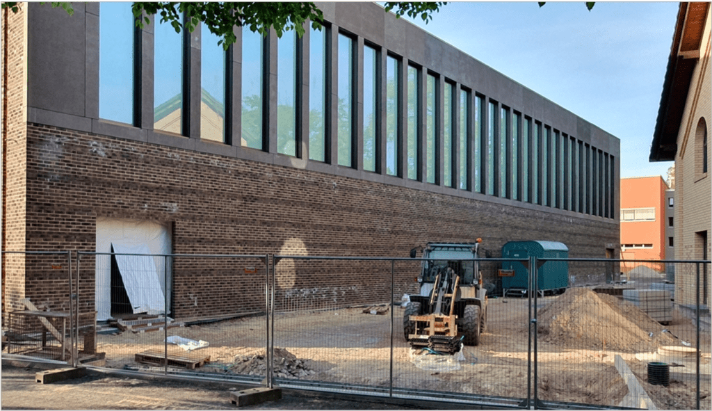 Eine Baustelle mit einem langen Backsteingebäude und hohen Fenstern. Innerhalb des eingezäunten Bereichs befinden sich ein Baggerlader und zwei mobile Toiletten. Auf dem Boden liegen verstreut Baumaterialien.