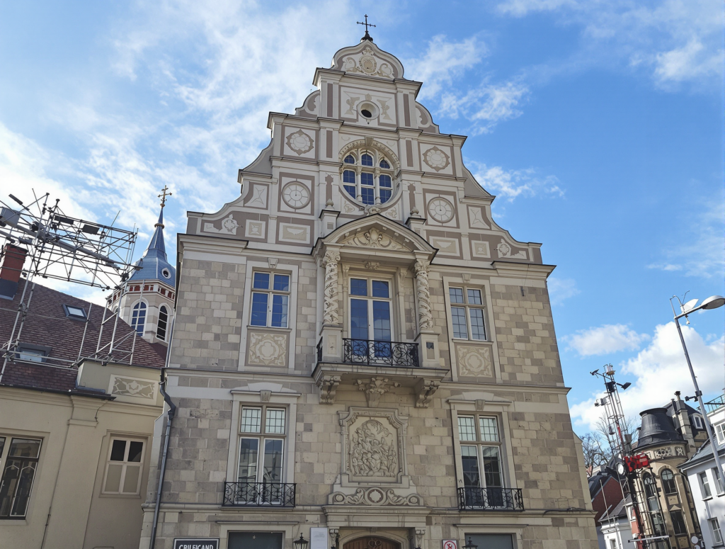 Ein kunstvolles, historisches Gebäude mit dekorativem Mauerwerk, Bogenfenstern und Balkonen steht unter einem blauen Himmel mit vereinzelten Wolken. Links sind angrenzende Dächer und ein Turm sichtbar.